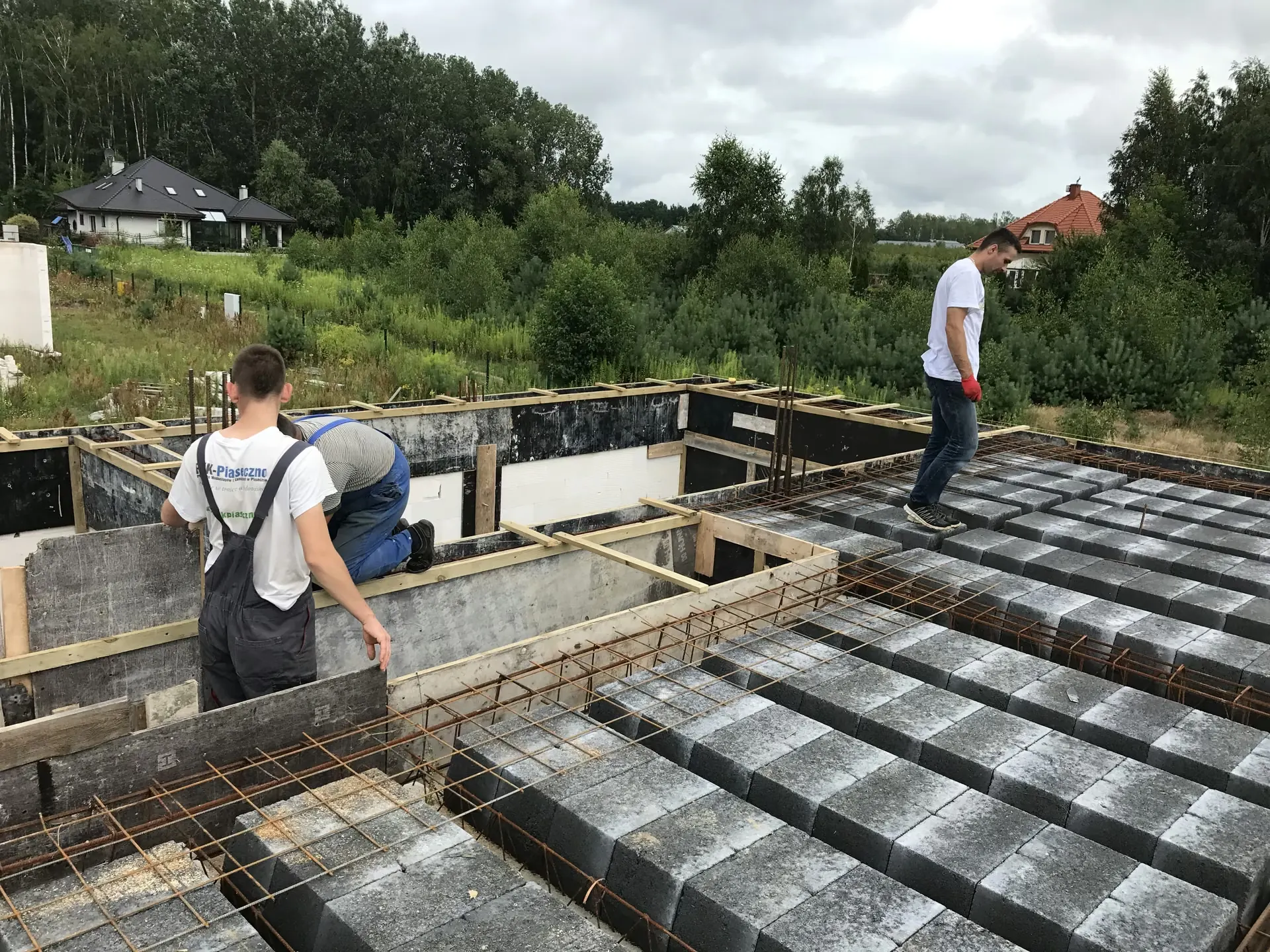 Trzech robotników budowlanych układa pustaki stropowe i zbrojenie nad płytą fundamentową domu jednorodzinnego, w tle dom sąsiada